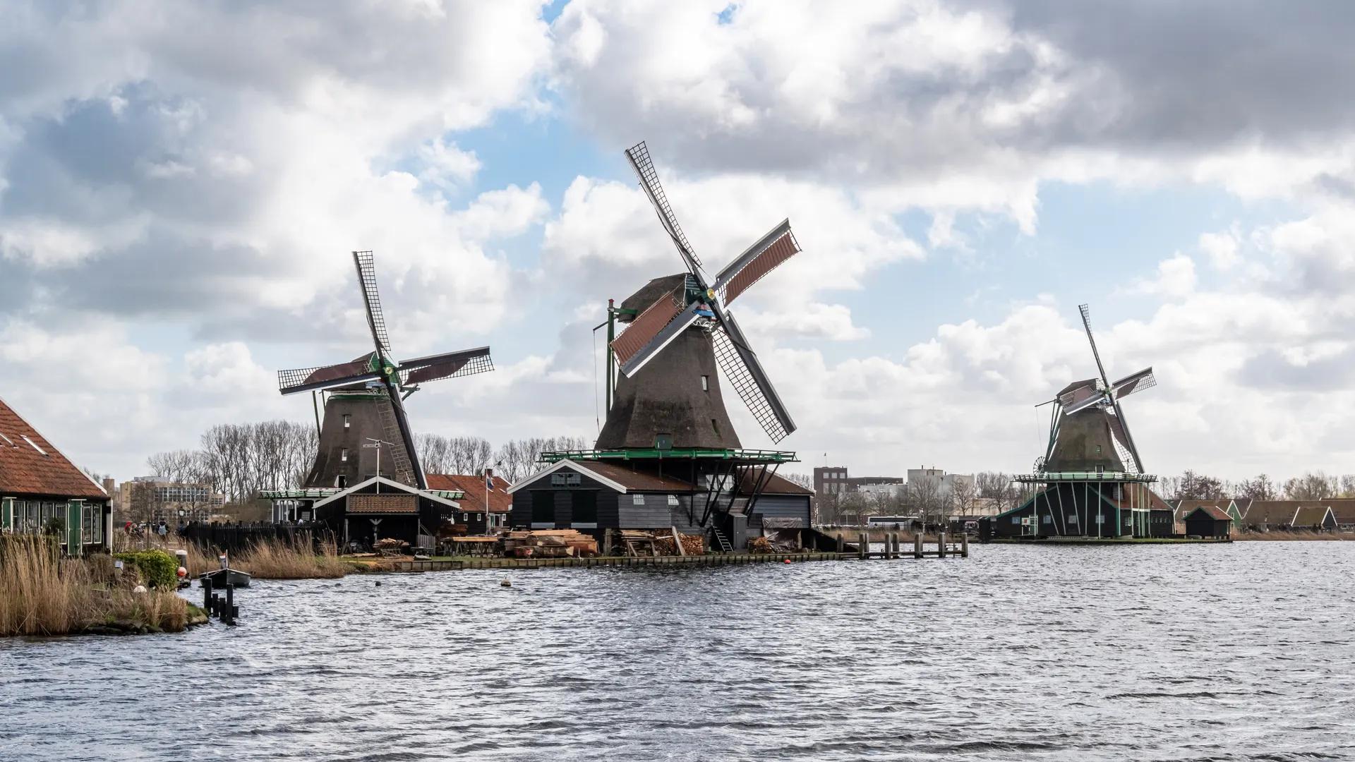 Three wooden windmills on the riverbank at Zaanse Schans under a cloudy sky